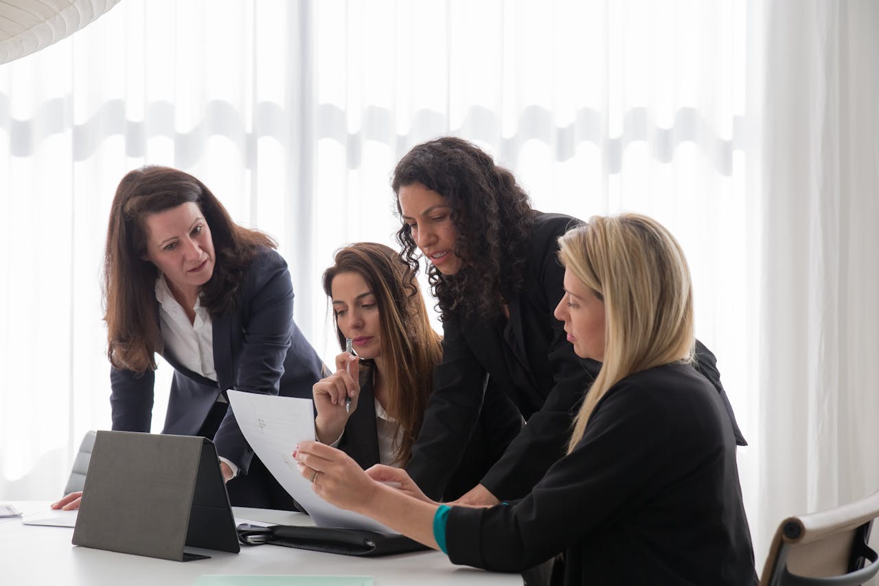 home-img Four businesswomen engaged in discussion over documents in a contemporary office setting.